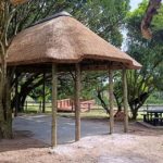 A freestanding outdoor entertainment lapa with a master-crafted thatch roof supported by sturdy timber poles, nestled under shaded garden trees