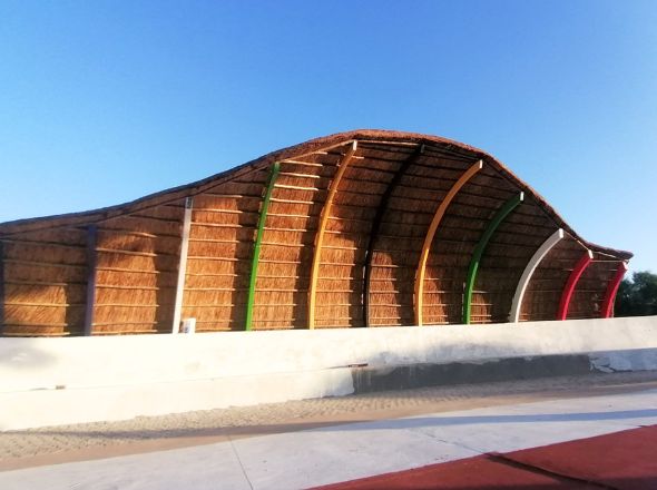 Projects An architecturally curved wave thatch canopy with colorful exposed support arches set over a paved outdoor stage.